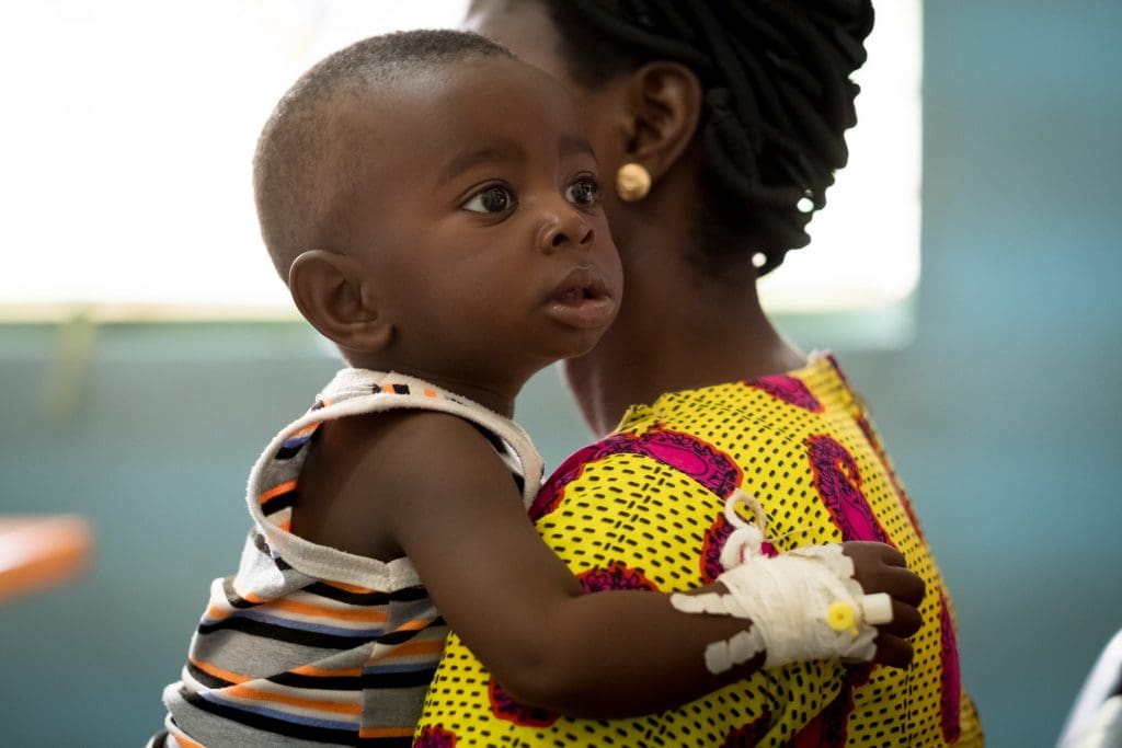 Kvinna som bär på ett barn som har en venkateter på handen.