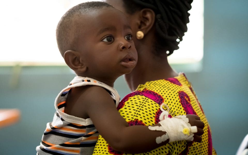 Kvinna som bär på ett barn som har en venkateter på handen.