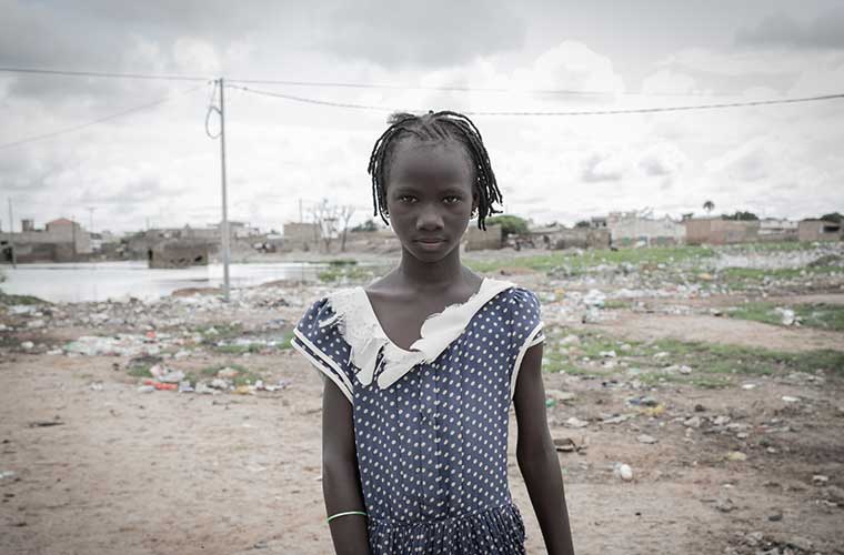 Allvarlig flicka i blå klänning står utomhus framför sandvidder.