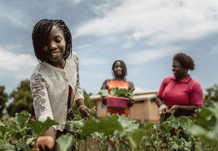 Ett barn ler och plockar salladsblad i SOS barnby i Moçambique. Två kvinnor syns i bakgrunden.