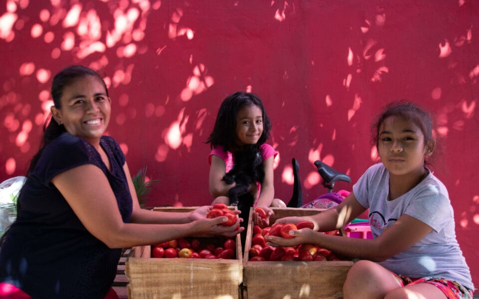 Kvinna från SOS Barnbyars familjestärkande program i Bolivia sitter med sina två döttrar vid två grönsakslådor.