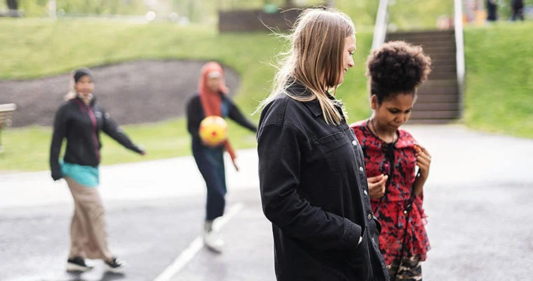 Två personer i förgrunden går och pratar. Två personer i bakgrunden spelar fotboll. De befinner sig utomhus.