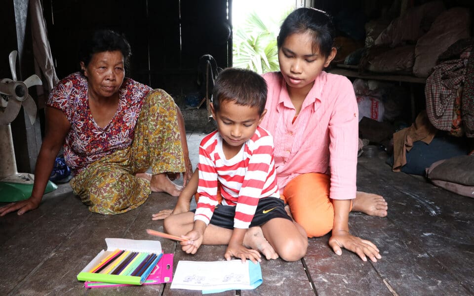 två kambodjanska barn sitter på golvet och ritar. Deras mamma sitter bakom dem.