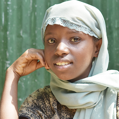 Flicka med huvudduk ler och har handen mot ansiktet står framför grön plåtvägg