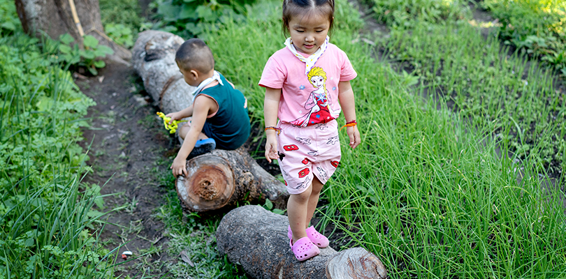 Litet barn går på en stock i rosa foppatofflor bakom en pojke med gul dinosaurie