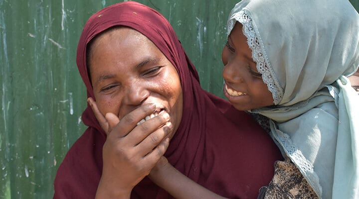 En mamma och dotter med huvudduk skrattar och kramas tillsammans mot grön vägg