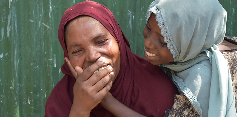 En mamma och dotter med huvudduk skrattar och kramas tillsammans mot grön vägg