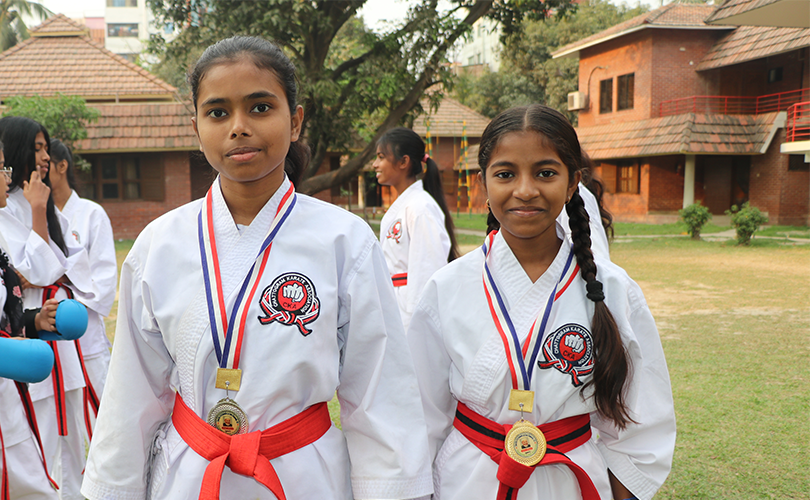 Två flickor i karatedräkt tittar in i kameran med gräs människor och hus bakom