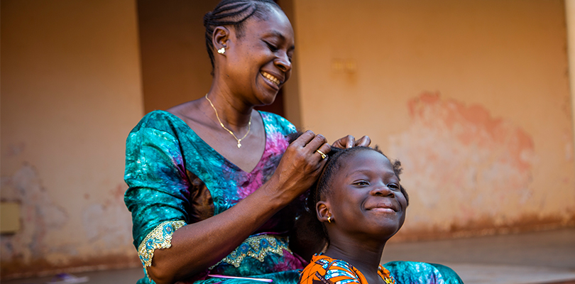 En mamma flätar barnets hår och båda ler utomhus mot orange bakgrund