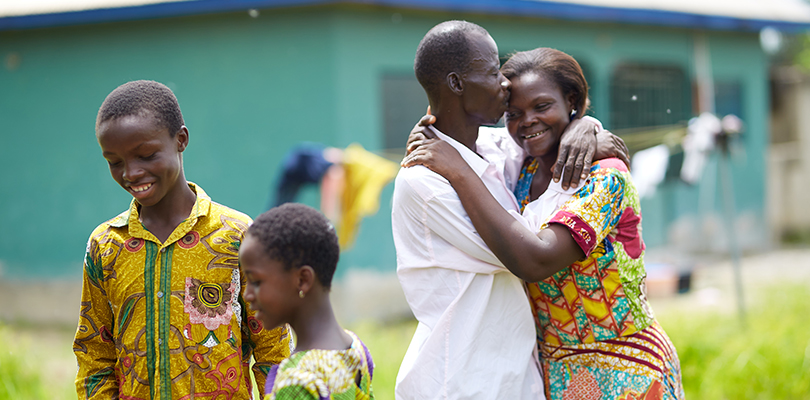 Två vuxna som omfamnar varandra med två barn intill på en solig gräsplätt med ett grönt hus i bakgrunden