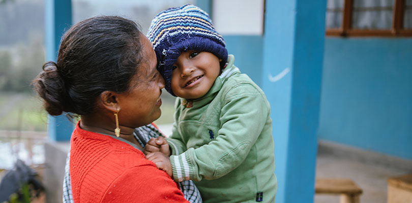 Kvinna i rött håller litet barn i grön jacka utomhus mot blå vägg