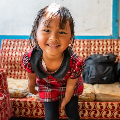 Glad flicka med busig blick framför en rödbrokig soffa tittar i kameran