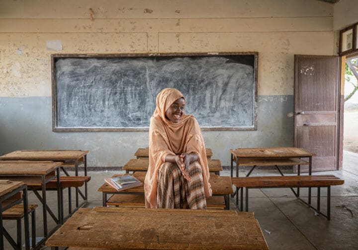 Flicka i orange huvudduk och klänning sitter på en bänk i ett klassrum