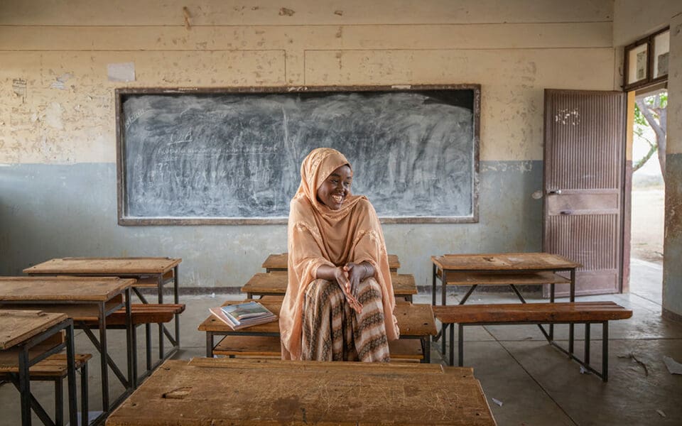 Flicka i orange huvudduk och klänning sitter på en bänk i ett klassrum