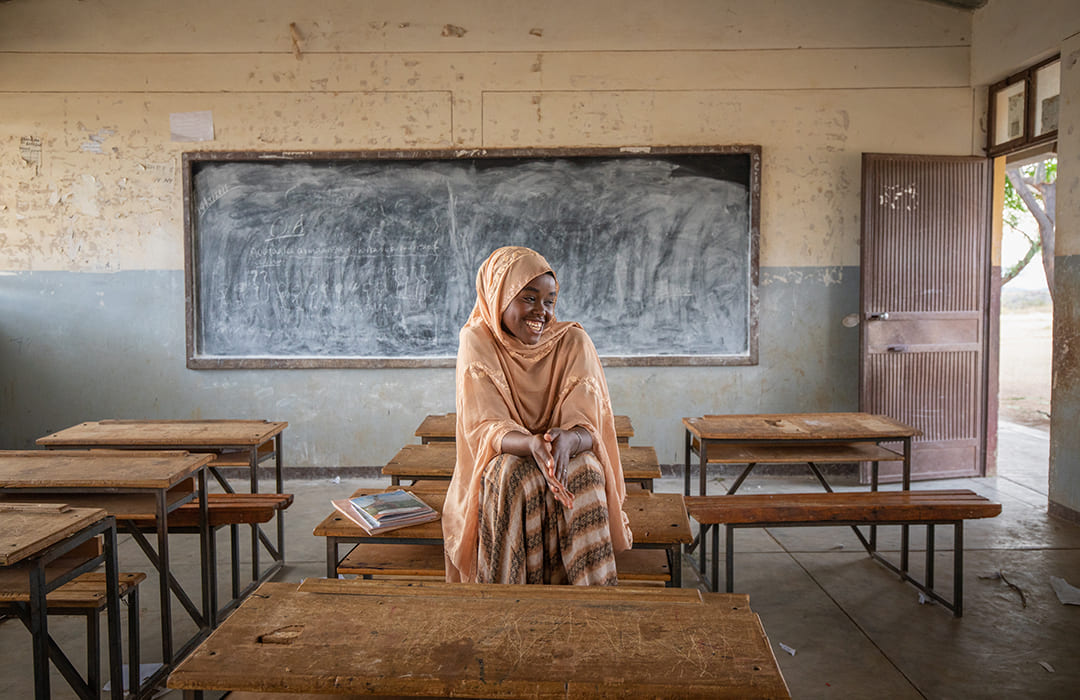 Flicka i orange huvudduk och klänning sitter på en bänk i ett klassrum